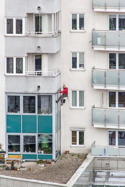 A window cleaner dressed in red safety gear is mid-task, using a harness to hold onto the exterior of a multi-storey residential building with white facade. The worker is cleaning the large glass wind