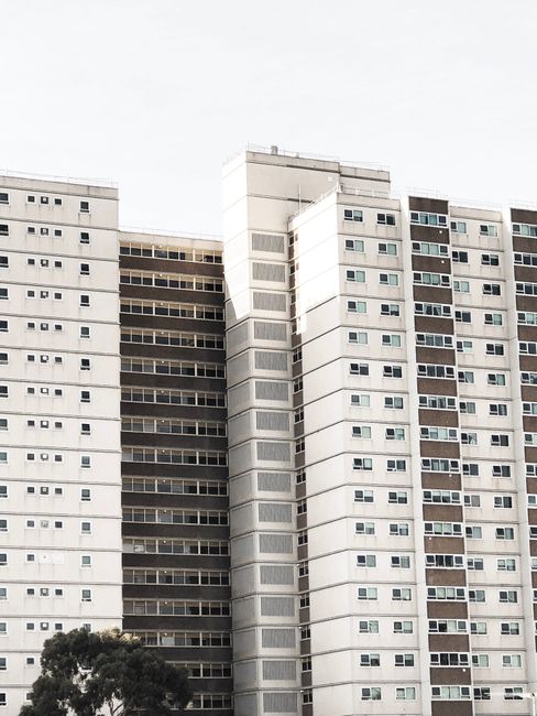 A window cleaner dressed in red safety gear is mid-task, using a harness to hold onto the exterior of a multi-storey residential building with white facade. The worker is cleaning the large glass wind