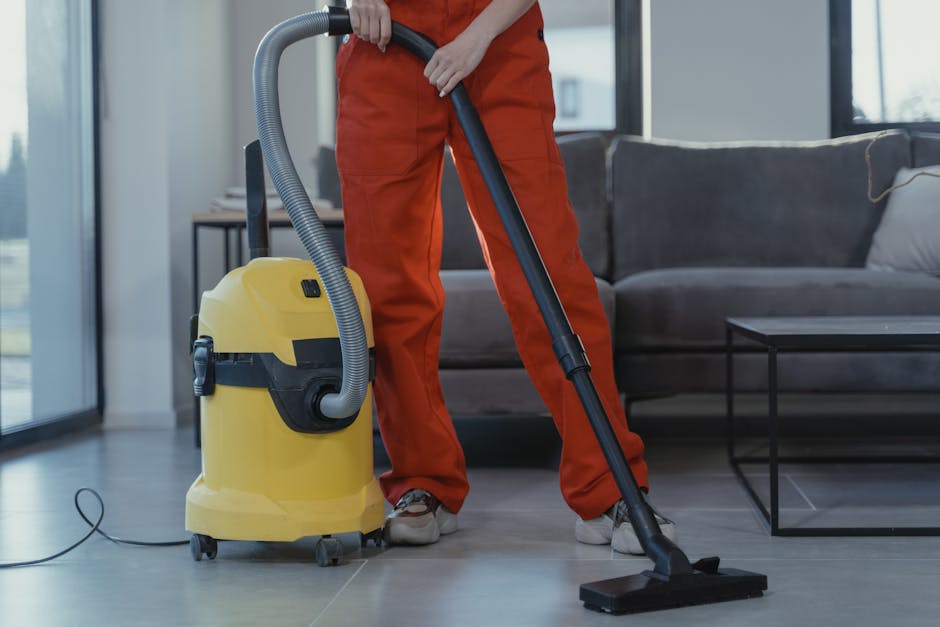 A professional cleaner from Carpet Cleaners NW5 operating a yellow wet/dry vacuum cleaner with a flexible hose, vacuuming a modern living room with a grey fabric sofa, a black metal coffee table, and large windows allowing natural light to illuminate the space. The cleaner is wearing orange work overalls and white shoes, focusing on surface cleaning of the tiled floor. The room appears clean and well-maintained, reflecting thorough deep cleaning practices typical of domestic cleaning services for landlords on Fortess Road NW5.
