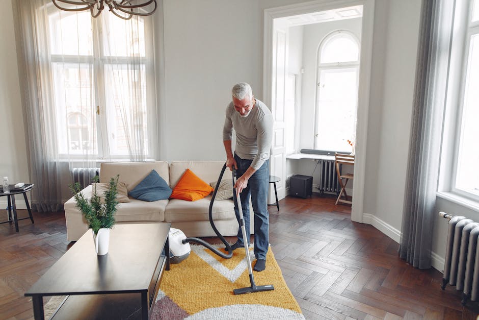 A middle-aged man with gray hair and a beard is performing deep cleaning on a wooden floor in a bright, modern living room using a vacuum cleaner. The room features large windows with sheer curtains, allowing natural light to illuminate the space. There is a white sofa with colorful cushions, a coffee table with a potted plant, and a yellow and white patterned rug on the floor. The walls are painted white, and the room contains minimal furniture, including a small side table and a desk with a chair near the window. The scene showcases a clean, well-maintained interior in the process of surface cleaning, highlighting attention to hygiene and maintenance, as provided by Carpet Cleaners NW5, specialists in domestic cleaning and surface sanitisation for landlords on Fortess Road NW5, NW5.