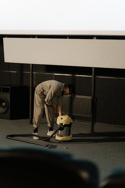 A professional cleaner from Carpet Cleaners NW5 operating a yellow wet/dry vacuum cleaner with a flexible hose, vacuuming a modern living room with a grey fabric sofa, a black metal coffee table, and large windows allowing natural light to illuminate the space. The cleaner is wearing orange work overalls and white shoes, focusing on surface cleaning of the tiled floor. The room appears clean and well-maintained, reflecting thorough deep cleaning practices typical of domestic cleaning services for landlords on Fortess Road NW5.