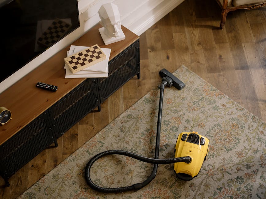 A yellow vacuum cleaner with a black hose and metal wand attachment is positioned on a floral patterned area rug in a living room corner. The hardwood floor is visible around the rug, and a black metal and wood media console is against the wall, holding a remote control, a stack of papers with a chessboard illustration, and a white decorative bust. The room is illuminated by natural light, highlighting the clean and tidy appearance of the space, which showcases surface cleaning and maintenance typical of residential environments managed by Carpet Cleaners NW5, in accordance with Camden Council carpet disposal rules.
