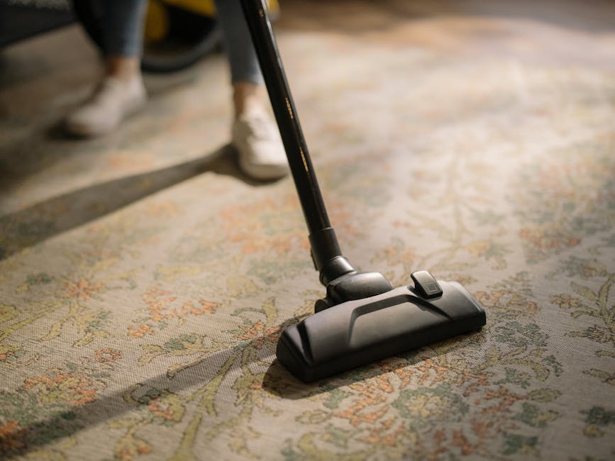 A close-up of a person vacuuming a patterned beige and green carpet in a room, with their legs and shoes visible in the background. The vacuum cleaner's black head and hose are shown in detail, with the surrounding carpet appearing clean and well-maintained under soft lighting. This image depicts surface cleaning using a modern vacuum to ensure hygiene and cleanliness, in line with domestic and commercial cleaning standards maintained by Carpet Cleaners NW5.
