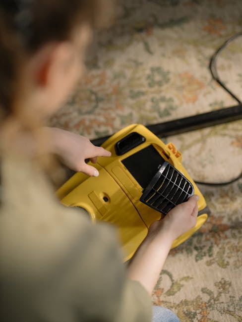 A yellow vacuum cleaner with a black hose and metal wand attachment is positioned on a floral patterned area rug in a living room corner. The hardwood floor is visible around the rug, and a black metal and wood media console is against the wall, holding a remote control, a stack of papers with a chessboard illustration, and a white decorative bust. The room is illuminated by natural light, highlighting the clean and tidy appearance of the space, which showcases surface cleaning and maintenance typical of residential environments managed by Carpet Cleaners NW5, in accordance with Camden Council carpet disposal rules.