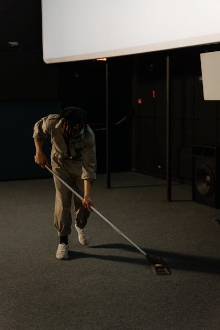 A person with short dark hair, wearing a red headband, pink shirt, and blue apron, is operating a vacuum cleaner on a patterned area rug in a living room. The room features a gray fabric sofa adorned