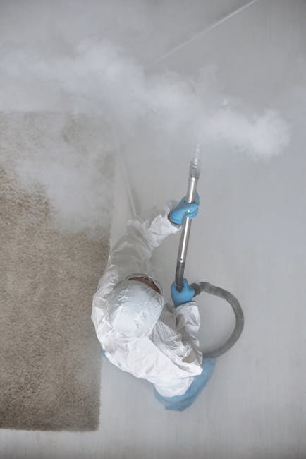A professional cleaner in protective white coveralls and blue gloves is using a steam or vapor cleaning tool on a white tiled surface in a residential setting, with visible steam or mist emanating from the device. The cleaning process appears to involve sanitisation or deep cleaning of the tiled floor or wall area. The surrounding environment includes a beige carpet and a section of smooth, clean white wall, illuminated by natural or overhead lighting. The scene is indicative of a thorough, hygienic cleaning operation, likely conducted by Carpet Cleaners NW5 as part of their upholstery cleaning or surface sanitisation services in Kentish Town Road, NW5, in line with their expertise in detailed domestic or commercial cleaning.