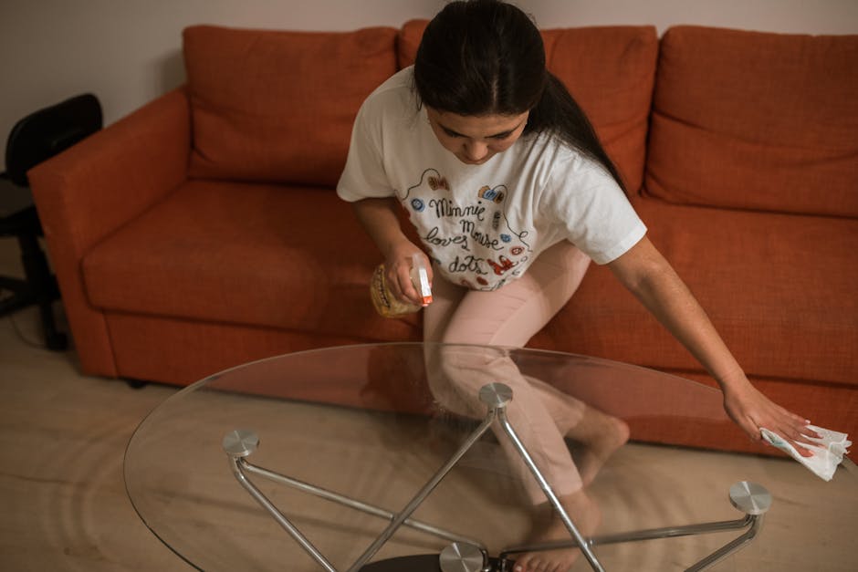 A woman with dark hair, wearing a white T-shirt with colorful text and beige pants, is kneeling on the floor and cleaning a glass coffee table with a cloth in her right hand, while holding a spray bottle in her left hand. The transparent glass table has a round surface supported by metal legs and is situated in a living room with a brown fabric sofa in the background. The room is well-lit, creating a clean and tidy appearance, with the surface of the table appearing spotless and reflective, emphasizing surface cleaning and sanitisation. The setting reflects domestic cleaning activities, highlighting detailed surface cleaning and maintenance practices that Carpet Cleaners NW5 offers.