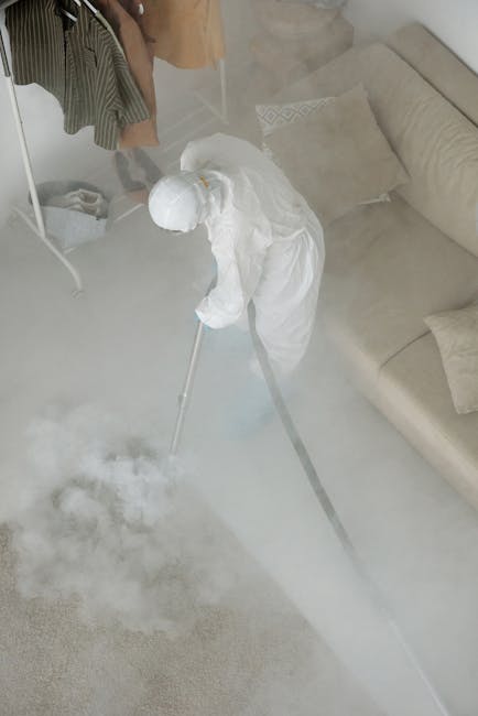 A professional cleaner in protective white coveralls and blue gloves is using a steam or vapor cleaning tool on a white tiled surface in a residential setting, with visible steam or mist emanating from the device. The cleaning process appears to involve sanitisation or deep cleaning of the tiled floor or wall area. The surrounding environment includes a beige carpet and a section of smooth, clean white wall, illuminated by natural or overhead lighting. The scene is indicative of a thorough, hygienic cleaning operation, likely conducted by Carpet Cleaners NW5 as part of their upholstery cleaning or surface sanitisation services in Kentish Town Road, NW5, in line with their expertise in detailed domestic or commercial cleaning.