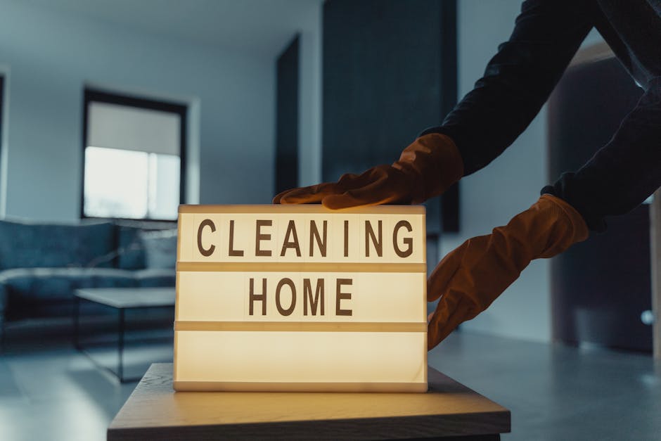 A person wearing brown rubber gloves is adjusting a decorative illuminated sign on a table, which reads 'CLEANING HOME' in bold black letters. The background features a modern living room with a blue sofa, a coffee table, a large window allowing natural light, and dark-colored walls, all appearing clean and well-maintained. The scene emphasizes domestic surface cleaning and deep cleaning practices associated with end of tenancy cleaning services in NW5, with a focus on hygiene and meticulous sanitisation by Carpet Cleaners NW5.