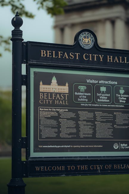 Close-up of an outdoor Belfast City Hall informational signboard mounted on a black metal post with decorative finials. The sign displays the Belfast City Hall logo, details about visitor attractions such as guided tours, self-guided tours, and the Bobbin Coffee Shop, along with bye-laws for city hall grounds. The background includes a blurred view of green trees and a cloudy sky, highlighting the outdoor setting. The sign is well-maintained and legible, reflecting the historical and civic importance of the location. For residential or commercial cleaning, such signage emphasizes the importance of surface cleaning and maintenance in public spaces, aligning with the expertise of Carpet Cleaners NW5 in deep cleaning and sanitisation practices.