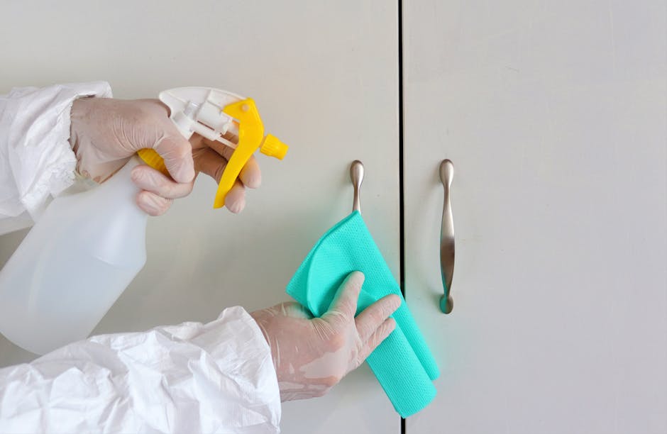 A person wearing white gloves and a protective disposable coverall is holding a white spray bottle with a yellow nozzle in one hand and a turquoise cloth in the other, cleaning the metal handles of white kitchen cabinet doors. The cabinet surfaces appear smooth and clean, with a slight reflection of ambient light. The background shows a plain, light-colored wall, emphasizing the focus on surface cleaning and sanitisation processes, which are part of a deep cleaning routine. The scene highlights tools used for domestic cleaning, along with a focus on maintaining hygiene in a kitchen environment, consistent with tips to avoid hidden cleaning costs in Kentish Town, NW5. The overall setting is well-lit, showcasing a tidy, hygienic space, and features the professional services of Carpet Cleaners NW5.