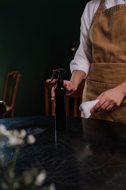 A person wearing a white long-sleeve shirt and a brown apron is holding a black spray bottle in one hand and a white cleaning cloth in the other, preparing for surface cleaning in a kitchen or dining area. The dark wooden table in front of them has a smooth, polished surface with some visible dust or residue, indicating an active cleaning process. In the background, there are wooden chairs with curved backs and a dark green wall, suggesting a cozy, well-maintained room. Lighting is soft and warm, highlighting the cleanliness and readiness for sanitisation or deep cleaning tasks, consistent with their professional cleaning service at Carpet Cleaners NW5 focusing on maintaining hygiene and cleanliness in residential or commercial spaces.