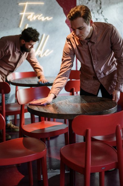 In a modern restaurant setting at The Forum Kentish Town, NW5, two staff members engaged in surface cleaning. The foreground shows a young man with short brown hair, wearing a light pink shirt, wiping down a round wooden table with a cloth, ensuring the surface is spotless and sanitized. The table has a dark, polished wooden top and is surrounded by red plastic chairs with curved backs, arranged neatly around the table. In the background, another staff member with dark curly hair and a beard, also dressed in a pink shirt, is wiping a similar table, contributing to maintaining high standards of cleanliness. The restaurant features a concrete wall with artistic neon lighting and some decorative elements, reflecting a contemporary interior design. The lighting is warm, creating a comfortable atmosphere, and the overall scene emphasizes thorough surface cleaning and hygiene practices as part of professional domestic and commercial cleaning services provided by Carpet Cleaners NW5. The focus is on ensuring tidy, well-maintained dining areas through proper sanitisation procedures.