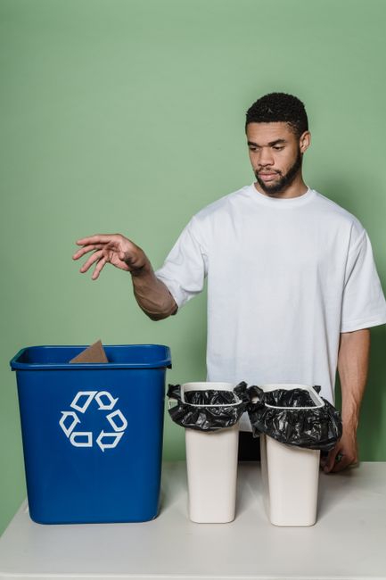 Two waste disposal bins placed against a plain beige wall in an indoor setting. The bin on the left is green with a black lid, designated for recycling, and features signage with a recycling symbol an