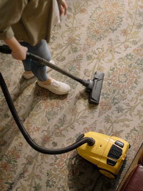 A person wearing beige shoes, blue jeans, and a tan coat vacuuming a patterned area rug in a residential room. The vacuum cleaner is yellow with a black hose and a push-button control, positioned on the carpet and connected to the cleaning head being used on the surface. The room features a floral or decorative carpet with light and muted tones, and the lighting suggests a well-lit indoor environment. The scene reflects surface cleaning, focusing on maintaining hygienic and deep cleaning standards, consistent with services offered by Carpet Cleaners NW5 in Kentish Town, NW5.