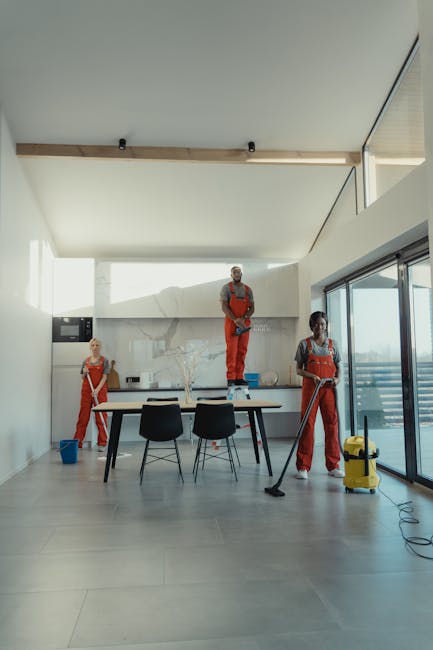 A professional cleaner from Carpet Cleaners NW5 is performing surface cleaning in a modern kitchen and dining area. The cleaner is dressed in orange overalls and using a mop on the tiled floor, which appears clean and well-maintained. In the background, a second cleaner in similar attire is seen near a large window with natural light illuminating the space. The kitchen has a minimalist design with a marble backsplash, a wooden dining table, and black chairs. The room features large glass sliding doors, allowing for ample daylight, and a blue cleaning bucket is positioned near the cleaner. The overall environment appears tidy, with surfaces free of dust and dirt, demonstrating effective deep cleaning and sanitisation for domestic or commercial spaces, consistent with the services offered by Carpet Cleaners NW5.